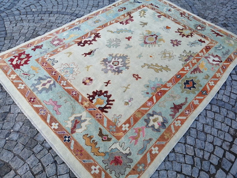 May include: A rectangular area rug with a cream-colored central field and a detailed border. The border features a light blue band with floral and geometric patterns, framed by a rust-colored band. The rug's design includes various colorful floral motifs.