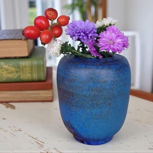 Könnte beinhalten: Eine blaue Vase mit strukturierter Oberfläche enthält einen Strauß aus lila und weißen Blumen und roten Beeren. Die Vase steht auf einem weißen Tisch neben einem Stapel alter Bücher mit grünen, braunen und grauen Einbänden.