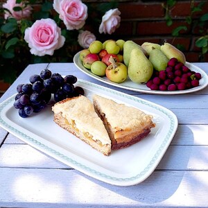 Op de afbeelding: Een stilleven met een witte rechthoekige schaal met twee stukken cake. Een tros donkere druiven en een schaal met fruit, waaronder peren, appels en druiven, zijn ook aanwezig. Roze rozen en een bakstenen muur zijn op de achtergrond te zien.