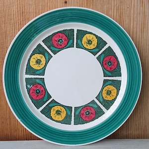 Op de afbeelding: Een wit keramiek bord met een groene rand en een herhalend patroon van gestileerde bloemen in rood, geel en groen.