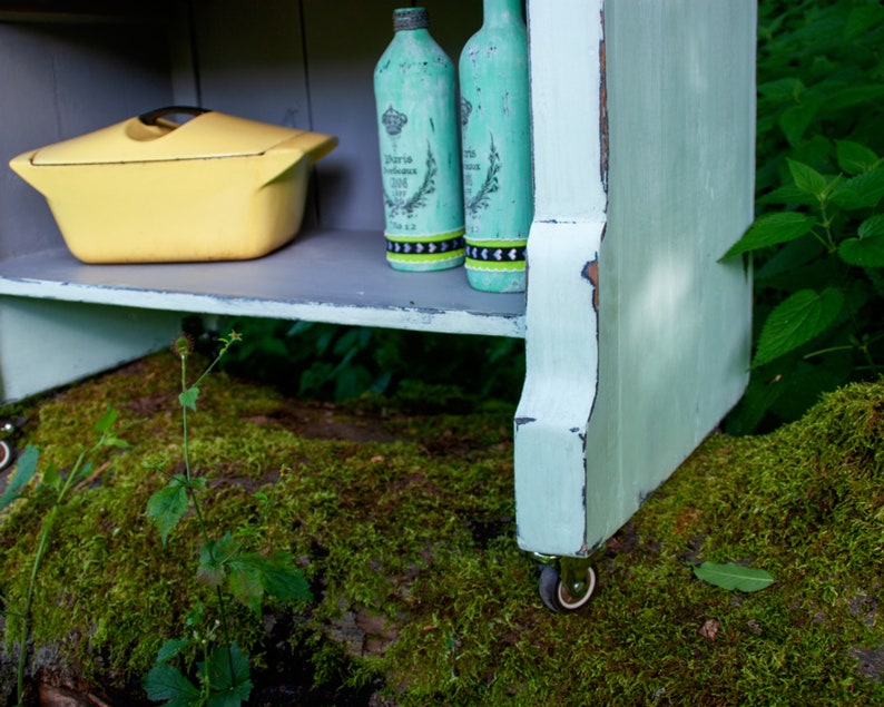 May include: A light blue wooden shelf unit with two shelves, a yellow ceramic casserole dish, and two green glass bottles with black and white labels. The shelf unit is on wheels and is sitting on a bed of moss.