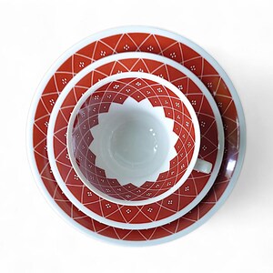 K&ouml;nnte beinhalten: Vintage Teeservice mit rotem und wei&szlig;em geometrischem Muster. Das Set besteht aus einer Teetasse, einer Untertasse und einem Teller. Die Artikel sind auf einer wei&szlig;en Oberfl&auml;che angeordnet.