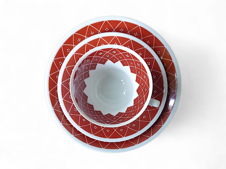 K&ouml;nnte beinhalten: Vintage Teeservice mit rotem und wei&szlig;em geometrischem Muster. Das Set besteht aus einer Teetasse, einer Untertasse und einem Teller. Die Artikel sind auf einer wei&szlig;en Oberfl&auml;che angeordnet.
