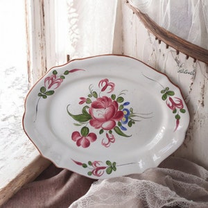 Może przedstawiać: Biała owalna ceramiczna tacka do serwowania z ręcznie malowanym wzorem kwiatowym. Wzór przedstawia różową różę z zielonymi liśćmi i różowymi i niebieskimi kwiatami.
