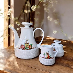 Op de afbeelding: Een witte keramische theeservies met bloemmotieven. De set bevat een theepot, een melkkan en een suikerpot. De theepot heeft een hoge, slanke vorm met een gebogen tuit en handvat.