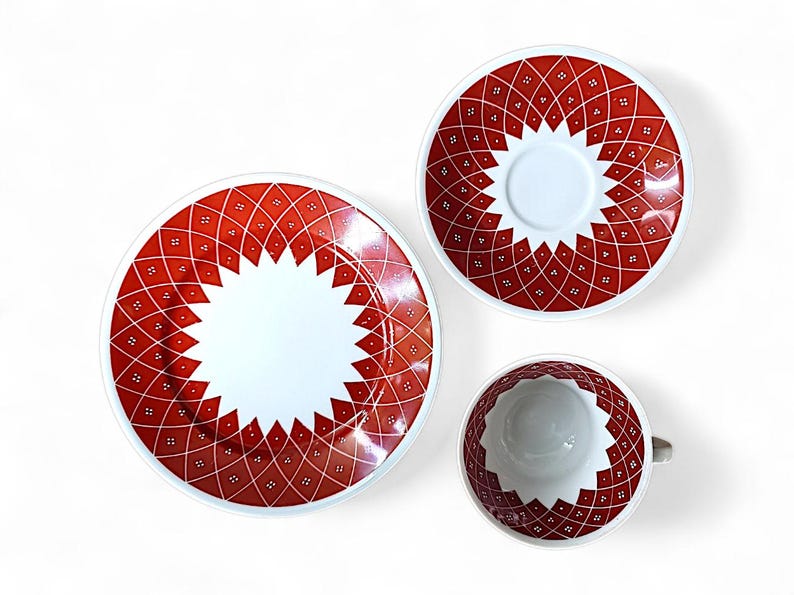 K&ouml;nnte beinhalten: Ein dreiteiliges Keramikgeschirr-Set mit einem rot-wei&szlig;en geometrischen Muster. Das Set umfasst einen gro&szlig;en Teller, eine Untertasse und eine Teetasse. Das Design hat ein sternf&ouml;rmiges Muster in der Mitte.