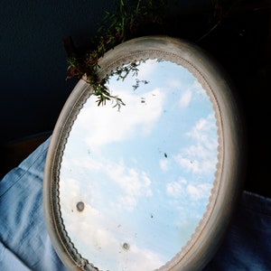 May include: An oval-shaped mirror with a white painted frame and a decorative lace trim. The mirror reflects a blue sky with white clouds.