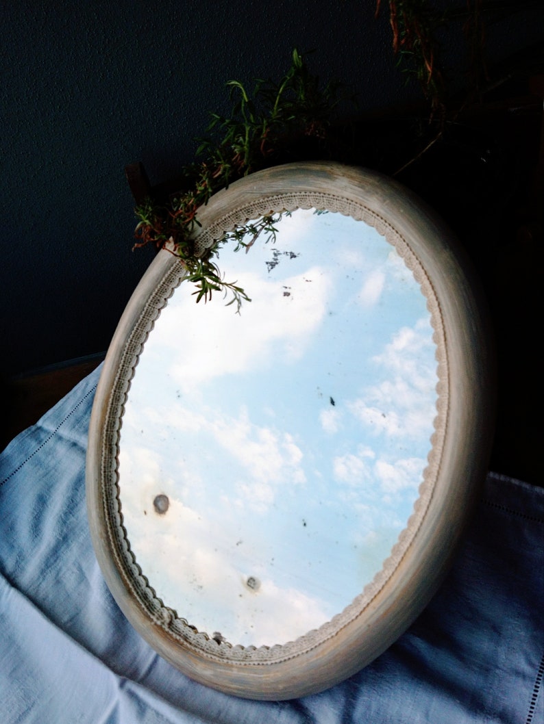 May include: An oval-shaped mirror with a white painted frame and a decorative lace trim. The mirror reflects a blue sky with white clouds.