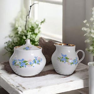 Könnte beinhalten: Ein weißes Porzellan-Zucker- und Milchkännchen-Set mit einem zarten Blumendesign mit blauen Vergissmeinnicht. Das Set hat einen goldenen Rand.