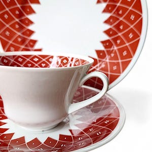K&ouml;nnte beinhalten: Wei&szlig;es Teeservice mit Tasse, Untertasse und Teller mit rotem geometrischem Muster. Die Tasse hat einen gebogenen Henkel und ein passendes Muster im Inneren. Untertasse und Teller haben ein &auml;hnliches Design.