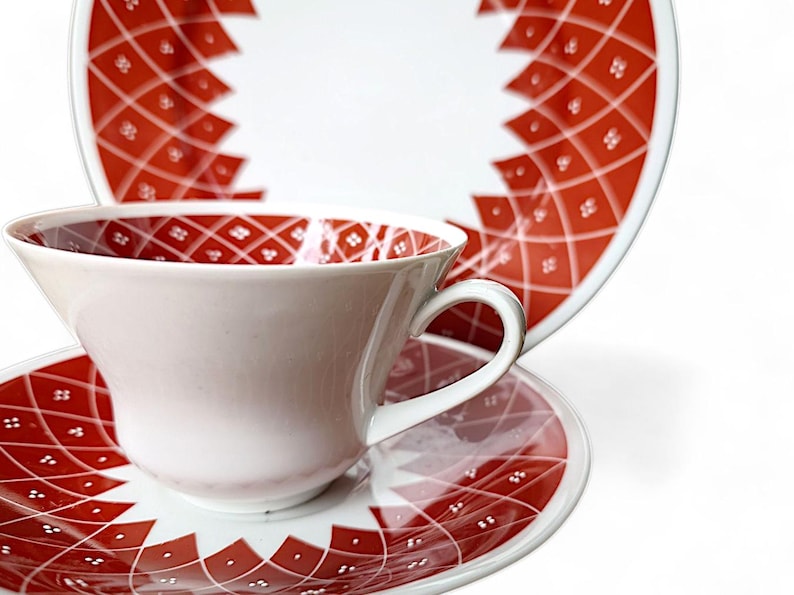K&ouml;nnte beinhalten: Wei&szlig;es Teeservice mit Tasse, Untertasse und Teller mit rotem geometrischem Muster. Die Tasse hat einen gebogenen Henkel und ein passendes Muster im Inneren. Untertasse und Teller haben ein &auml;hnliches Design.