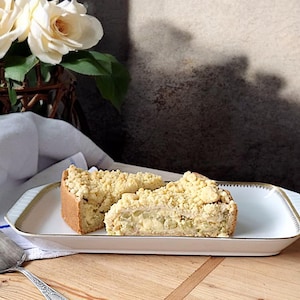 Könnte beinhalten: Zwei Scheiben eines goldbraunen Desserts mit Streuseln auf einem weißen rechteckigen Teller mit Goldrand. Ein silberner Tortenheber liegt auf einer Holzoberfläche. Ein Strauß weißer Rosen im Hintergrund.