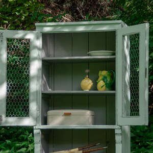 May include: A white cabinet with wire mesh doors and shelves. The cabinet has a light green paint finish and is filled with various items, including a plate, a bread box, and two ceramic lemons.