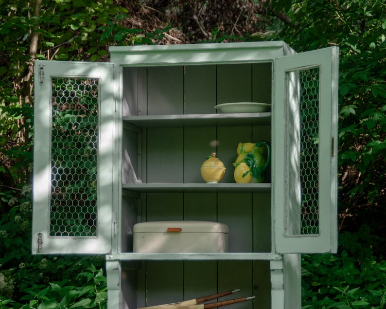 May include: A white cabinet with wire mesh doors and shelves. The cabinet has a light green paint finish and is filled with various items, including a plate, a bread box, and two ceramic lemons.