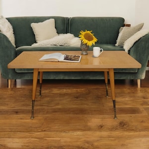 Könnte beinhalten: Ein heller Holz-Couchtisch mit einer rechteckigen Tischplatte und vier Beinen. Auf dem Tisch liegen ein Buch und eine Brille. Eine Vase mit einer Sonnenblume steht auf dem Tisch. Der Tisch steht vor einem grünen Sofa mit weißen Kissen.