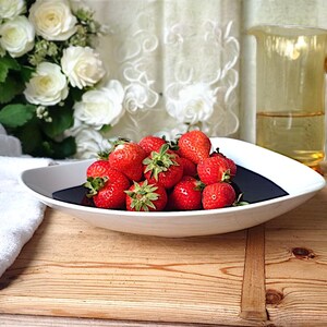 Op de afbeelding: Een witte en zwarte decoratieve kom gevuld met verse rode aardbeien. Een gele glazen kan met een gebogen handvat staat op de achtergrond, samen met een boeket witte rozen. De scène speelt zich af op een houten oppervlak.