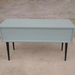 Das Bild zeigt eine original Vintage Kommode von hinten. Der gesamte Korpus, einschließlich des Rückens, ist in einem beruhigenden Grün gestrichen. Die langen, eleganten Holzbeine sind in Schwarz gehalten. Ein Blickfang mit Retro-Charme.