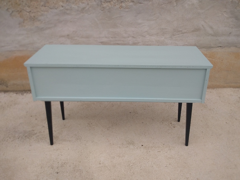 Das Bild zeigt eine original Vintage Kommode von hinten. Der gesamte Korpus, einschließlich des Rückens, ist in einem beruhigenden Grün gestrichen. Die langen, eleganten Holzbeine sind in Schwarz gehalten. Ein Blickfang mit Retro-Charme.