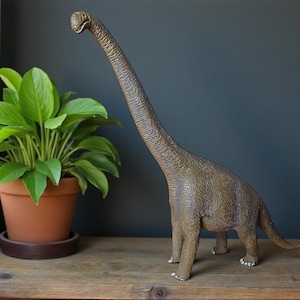 Könnte beinhalten: Eine detaillierte, braune Brachiosaurus-Dinosaurierfigur steht auf einer Holzoberfläche. Der langhalsige Dinosaurier hat eine strukturierte Haut und befindet sich in der Nähe einer Topfpflanze mit grünen Blättern. Der Hintergrund ist eine dunkelblaue Wand.