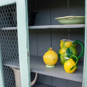 May include: A light green cabinet with a wire mesh door and gray shelves. The shelves are decorated with a lemon-themed pitcher, a sugar bowl, and a creamer.
