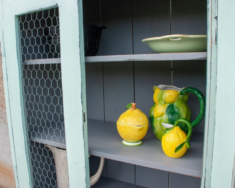 May include: A light green cabinet with a wire mesh door and gray shelves. The shelves are decorated with a lemon-themed pitcher, a sugar bowl, and a creamer.