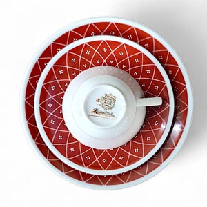 K&ouml;nnte beinhalten: Draufsicht auf ein Vintage-Teeservice. Das Set umfasst eine Teetasse, eine Untertasse und zwei Teller, alle mit einem rot-wei&szlig;en Rautenmuster. Die Tasse und Untertasse sind wei&szlig; mit einem goldenen Emblem. Die Teller haben einen Durchmesser von ca. 23 cm.