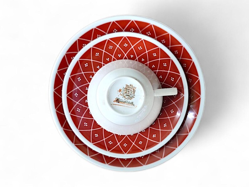 K&ouml;nnte beinhalten: Draufsicht auf ein Vintage-Teeservice. Das Set umfasst eine Teetasse, eine Untertasse und zwei Teller, alle mit einem rot-wei&szlig;en Rautenmuster. Die Tasse und Untertasse sind wei&szlig; mit einem goldenen Emblem. Die Teller haben einen Durchmesser von ca. 23 cm.