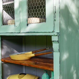 May include: A light green wooden cabinet with two glass doors and shelves. The doors have wire mesh inserts and the shelves are made of wood. There are two yellow pans and a wooden utensil on the top shelf.