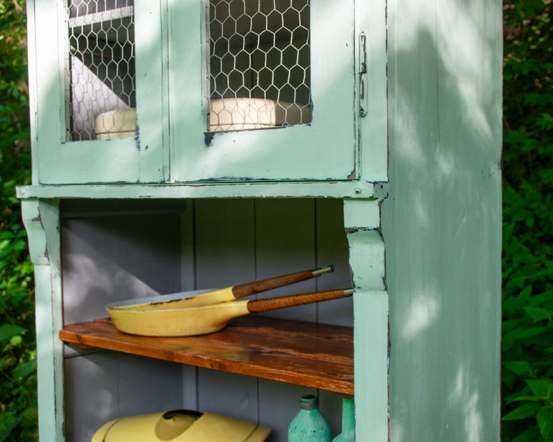 May include: A light green wooden cabinet with two glass doors and shelves. The doors have wire mesh inserts and the shelves are made of wood. There are two yellow pans and a wooden utensil on the top shelf.
