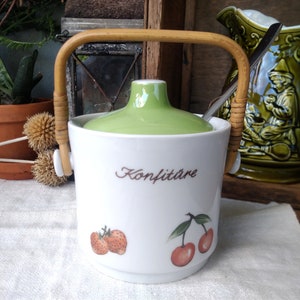 Pode incluir: Um pote de cerâmica branco com tampa verde e alça de vime. O pote tem um desenho floral com morangos e cerejas e a palavra "Konfitüre" escrita na frente.
