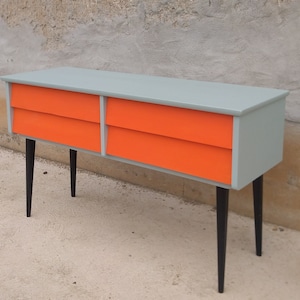 Das Bild zeigt eine original Vintage Kommode mit vier orange gestrichenen Schubladen. Der Korpus ist grün, und die langen Holzbeine sind schwarz gestrichen. Die Kommode wird leicht seitlich präsentiert, hat Retro-Flair