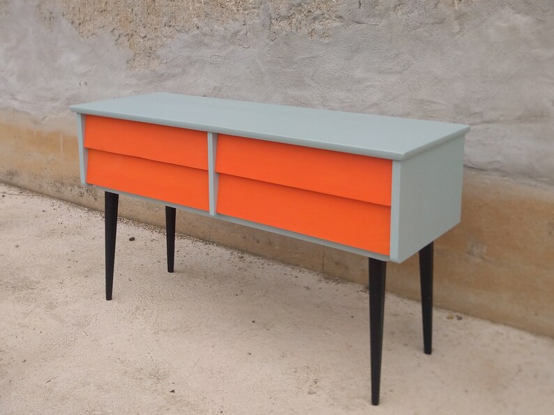 Das Bild zeigt eine original Vintage Kommode mit vier orange gestrichenen Schubladen. Der Korpus ist grün, und die langen Holzbeine sind schwarz gestrichen. Die Kommode wird leicht seitlich präsentiert, hat Retro-Flair