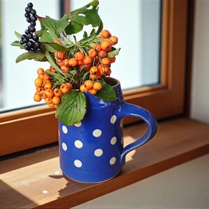 Op de afbeelding: Een blauwe keramische mok met witte stippen bevat oranje bessen en groene bladeren. De mok staat op een houten vensterbank. Het raam en het natuurlijke licht zijn op de achtergrond. Decoratief object.