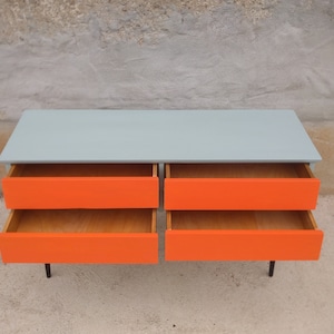 Das Bild zeigt eine original Vintage Kommode mit vier geöffneten Schubladen, die in Orange gestrichen sind. Der Korpus ist grün, und die langen Holzbeine sind schwarz gestrichen. Die frontale leicht von oben Präsentation betont den Retro-Charme