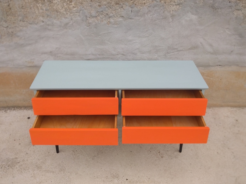 Das Bild zeigt eine original Vintage Kommode mit vier geöffneten Schubladen, die in Orange gestrichen sind. Der Korpus ist grün, und die langen Holzbeine sind schwarz gestrichen. Die frontale leicht von oben Präsentation betont den Retro-Charme