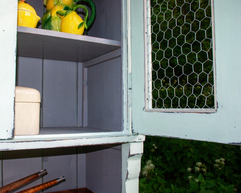 May include: A light blue cabinet with a wire mesh door. The cabinet has two shelves, one with a white box and the other with a ceramic lemon pitcher and two lemons.