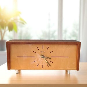 Peut inclure: Une horloge rectangulaire en bois avec un cadran en bois clair et un dessus et des côtés en bois foncé. Le cadran de l'horloge a un design minimaliste avec des lignes fines marquant les heures et des aiguilles dorées. L'horloge est soutenue par des pieds clairs et inclinés.