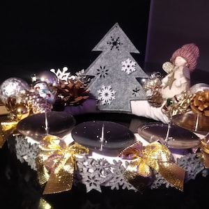 Könnte beinhalten: Ein weißer Adventskranz mit vier Kerzenhaltern, verziert mit goldenen Schleifen, silbernen Ornamenten, Tannenzapfen und einem grauen Filz-Weihnachtsbaum mit Schneeflocken. Eine Engelfigur mit rosa Flügeln und einem rosa Hut sitzt auf dem Kranz.