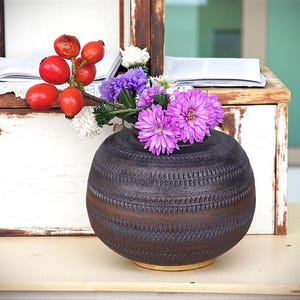 Könnte beinhalten: Eine dunkelbraune, runde Keramikvase mit einem strukturierten Muster, die lila und weiße Blumen enthält. Rote Beeren und ein Zweig mit Blumen sind in der Nähe der Vase platziert. Die Vase steht auf einer hellen Oberfläche.