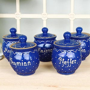 Può includere: Cinque barattoli di spezie in ceramica blu con coperchi. Ogni barattolo è decorato con pois bianchi e un'etichetta bianca. Le etichette riportano "Thymian", "Majora", "Pfeffer", "Muskat" e "Ka".