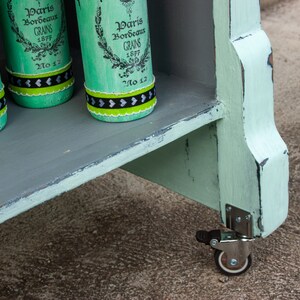 May include: A light green wooden shelf unit with three shelves and a distressed paint finish. The unit has four wheels and is decorated with four green glass bottles with black and white labels that read "Paris Bordeaux Grains 1877 No 12".