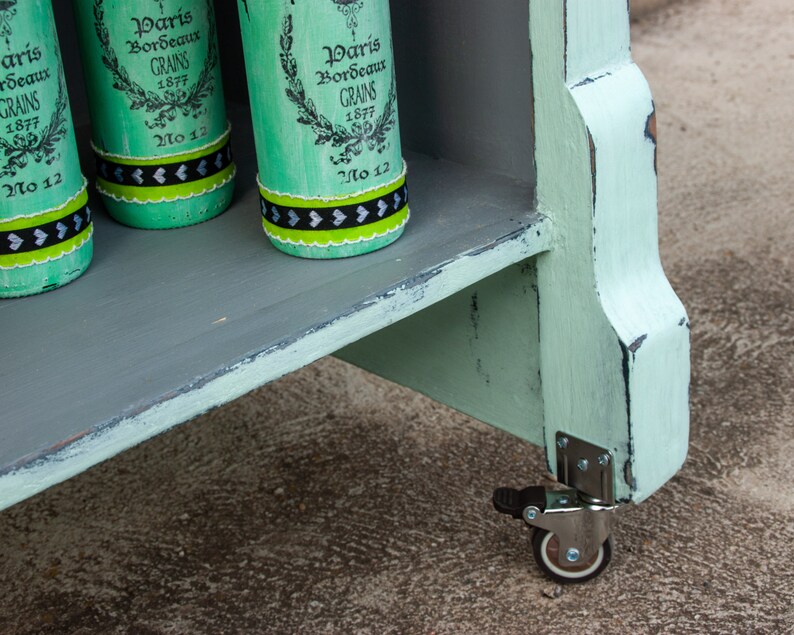 May include: A light green wooden shelf unit with three shelves and a distressed paint finish. The unit has four wheels and is decorated with four green glass bottles with black and white labels that read "Paris Bordeaux Grains 1877 No 12".
