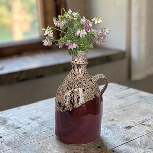 Könnte beinhalten: Eine Keramik-Krugvase mit zweifarbiger Glasur, bestehend aus einem dunkelroten Boden und einem gesprenkelten cremefarbenen Oberteil. Das Gefäß hat einen schmalen Hals, einen kleinen Henkel an der Seite und enthält kleine, zarte lila-weiße Wildblumen.