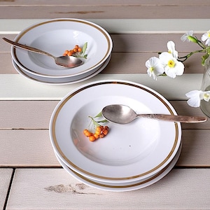 May include: White soup bowls with gold rims and silver spoons are arranged on a light-coloured wooden surface. Each bowl contains a sprig of orange berries. A glass vase with white flowers is in the background.