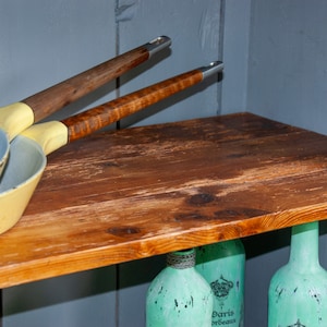 May include: Two yellow and blue frying pans with wooden handles hanging on a wooden shelf. The shelf is mounted on a gray wall. Three aqua blue glass bottles are visible below the shelf.