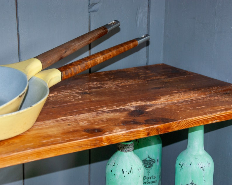 May include: Two yellow and blue frying pans with wooden handles hanging on a wooden shelf. The shelf is mounted on a gray wall. Three aqua blue glass bottles are visible below the shelf.