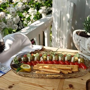 Può includere: Un vassoio di vetro con un assortimento di antipasti, tra cui olive, pomodorini, formaggio e grissini. Sono presenti anche un bicchiere di vino rosso e una fioriera decorativa a forma di conchiglia con piante. La scena è ambientata all'aperto su una superficie di legno.