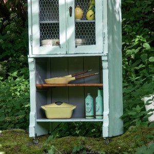 May include: A light blue painted cabinet with wire mesh doors and open shelves. The cabinet has a yellow casserole dish, wooden cooking utensils, and glass bottles on the shelves. The cabinet is on wheels and is in front of a green leafy background.