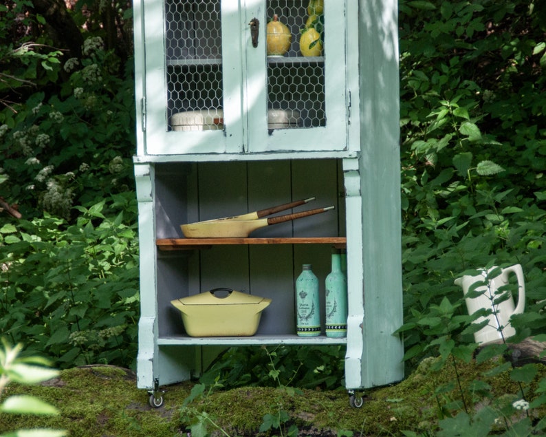 May include: A light blue painted cabinet with wire mesh doors and open shelves. The cabinet has a yellow casserole dish, wooden cooking utensils, and glass bottles on the shelves. The cabinet is on wheels and is in front of a green leafy background.