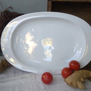 Könnte beinhalten: Eine weiße ovale Servierplatte mit einem erhöhten Rand. Drei rote Tomaten und ein Stück Ingwerwurzel liegen auf einer weißen Oberfläche vor der Platte.